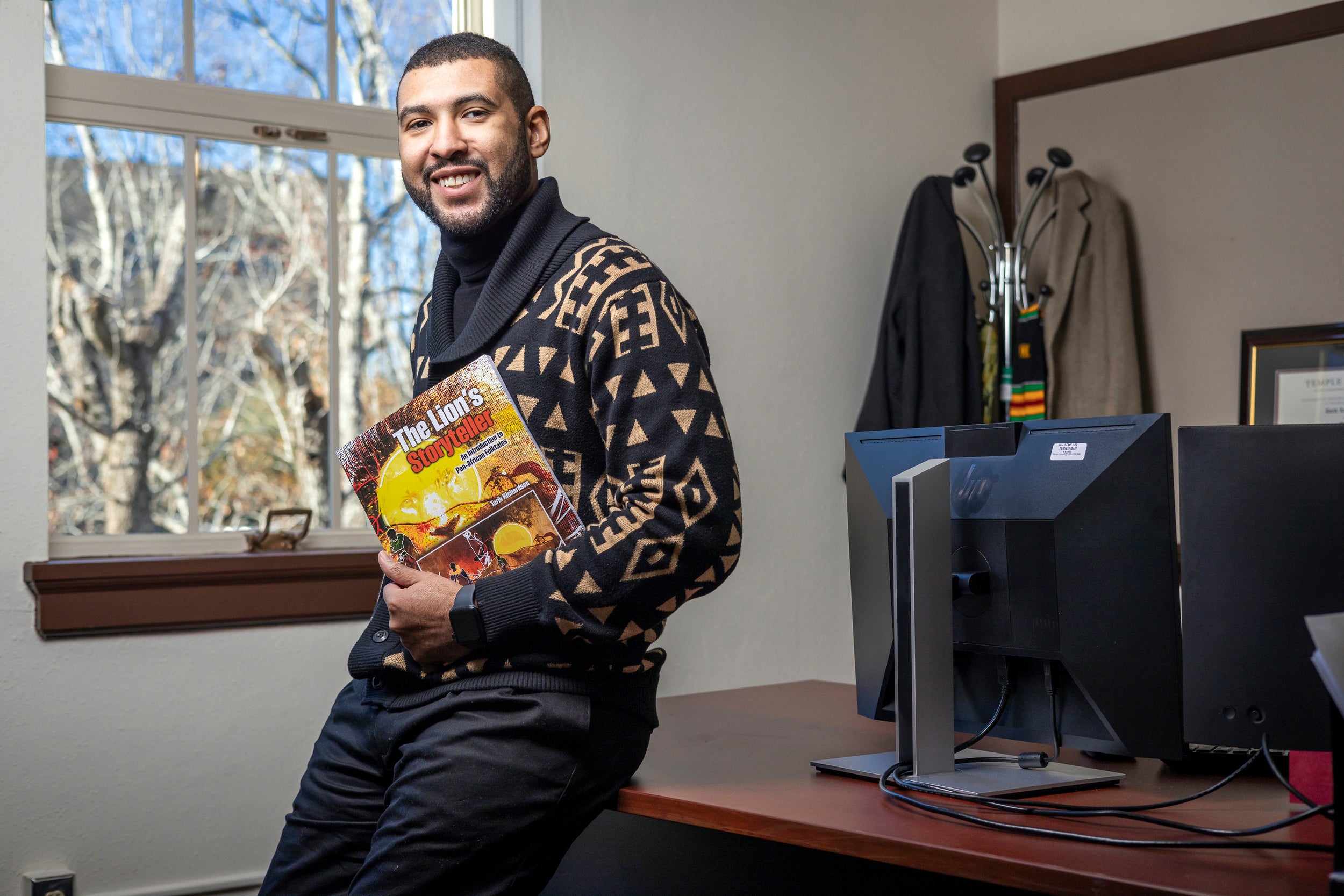 Picture of Tarik in an office holding a copy of his book: "The Lion's Storyteller: An Introduction to Pan-African Folktales"