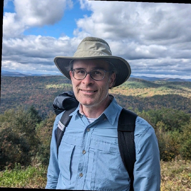 Portrait of Brent in hiking gear, smiling in front of a mountain landscape