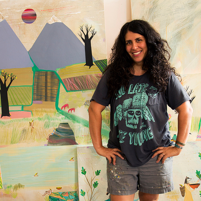 Deedee smiling and standing with both hands on her hips in front of several of her artwork pieces