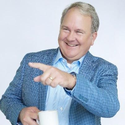 Michael has a mug in one hand and is smiling and pointing with his other hand. 
